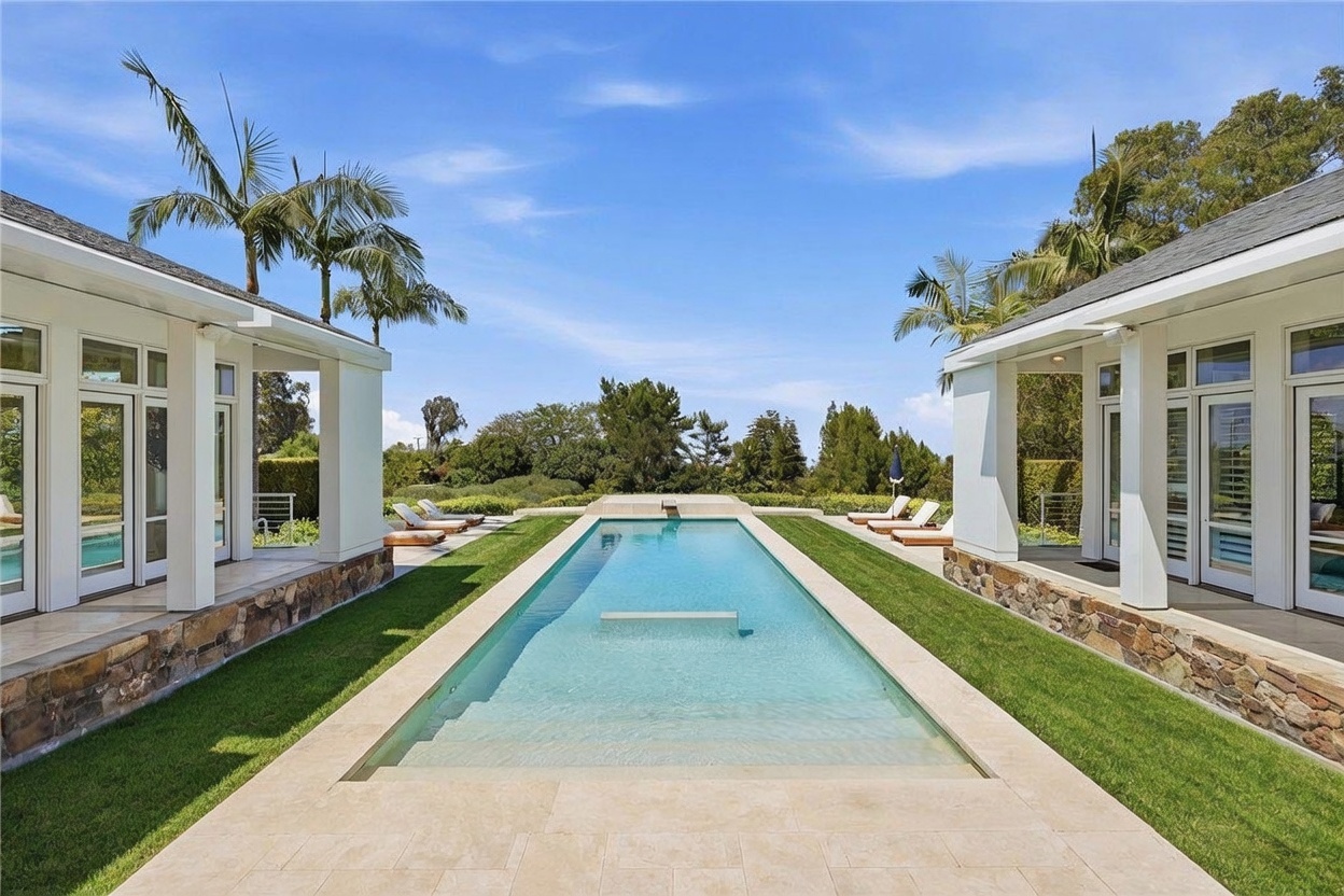 This symmetrical outdoor space features a long, rectangular swimming pool centered between two matching white structures with large glass doors. The pool is surrounded by manicured green lawns and light-colored stone decking, with lounge chairs positioned on either side. The perspective is a straight-on, eye-level shot that emphasizes the clean lines, architectural balance, and serene atmosphere of the property.