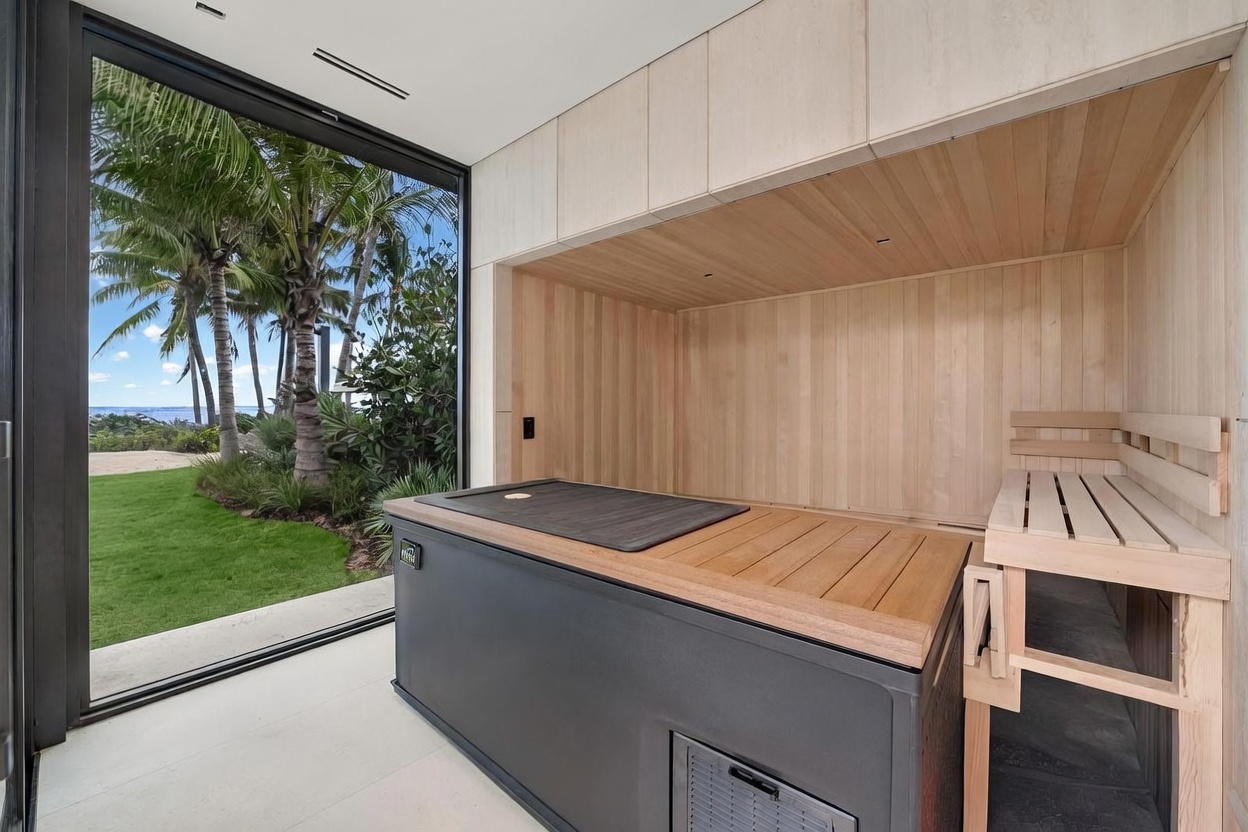 This interior space features a dedicated wellness area equipped with a large, dark-toned cold plunge or spa unit positioned against a light wood-paneled wall. To the right, a wooden bench provides seating, while a large glass wall offers an expansive view of a lush, tropical outdoor garden with palm trees. The room combines natural wood textures with modern, clean lines to create a serene and high-end spa-like atmosphere.
