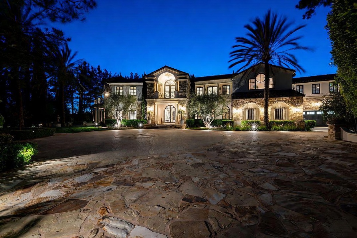 This grand estate is captured at twilight, showcasing a sprawling stone-paved motor court leading to an impressive multi-story residence. The architecture features a mix of stone and stucco finishes, with a prominent arched entryway and symmetrical window placement that creates a stately, formal appearance. Lush landscaping, including mature palm trees and manicured shrubs, is illuminated by warm exterior lighting, enhancing the home's curb appeal and luxurious atmosphere.