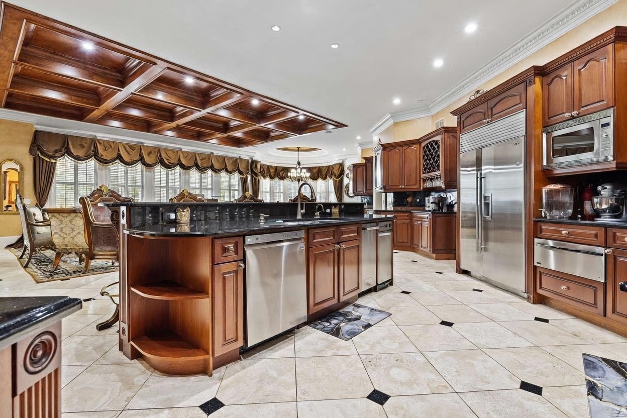 This expansive, traditional-style kitchen features a large central island with a dark stone countertop and integrated stainless steel appliances. The room is defined by its intricate coffered wood ceiling and light-colored tile flooring with dark accent inlays. A dining area is visible in the background, framed by large windows with elegant valances, creating a sophisticated and open atmosphere.