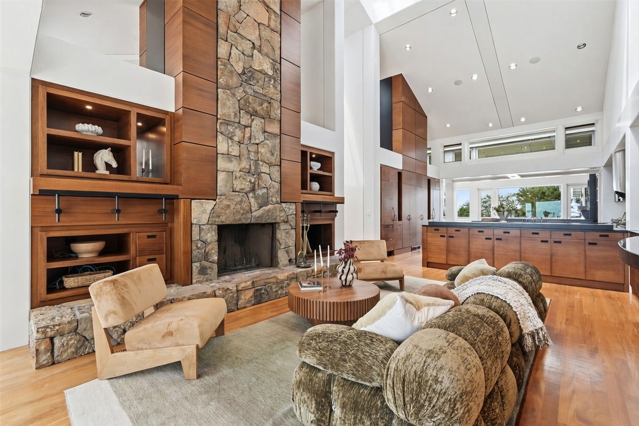 This expansive living room features a prominent stone-clad chimney breast that serves as a central focal point, flanked by custom wood cabinetry and shelving. The open-concept space includes a textured, rounded sofa, a circular wooden coffee table, and two modern lounge chairs set upon a large area rug. The room boasts high, vaulted ceilings and a seamless transition into an adjacent kitchen area, creating an airy and sophisticated atmosphere.