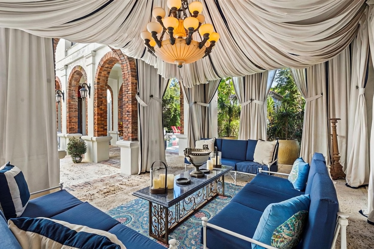This luxurious covered patio features an elegant draped fabric ceiling and a grand chandelier, creating a sophisticated outdoor living space. The area is furnished with plush blue sofas arranged around a central coffee table, set against a backdrop of classic brick arches and stone flooring. The open-air design provides a seamless transition to the lush garden and pool area beyond, offering a serene and upscale atmosphere.