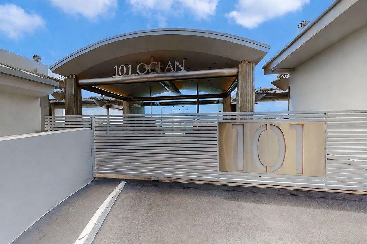 This image captures the modern, gated entrance to a property marked '101 Ocean.' A sleek, horizontal-slat metal gate features a prominent '101' plaque, leading to a covered entryway with a curved architectural roofline and glass doors. The perspective is a direct, eye-level shot that emphasizes the clean lines and contemporary aesthetic of the building's facade.