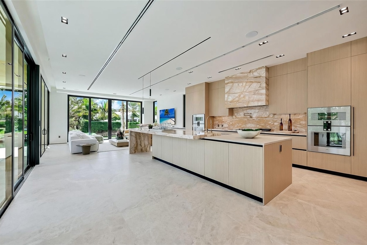 This expansive, open-concept kitchen features a large central island with a stone countertop and light wood cabinetry. A striking stone-clad backsplash and range hood serve as a focal point, complemented by integrated appliances and sleek, minimalist design elements. The space flows seamlessly into a bright living area, with large glass sliding doors providing an unobstructed view of the lush outdoor greenery.