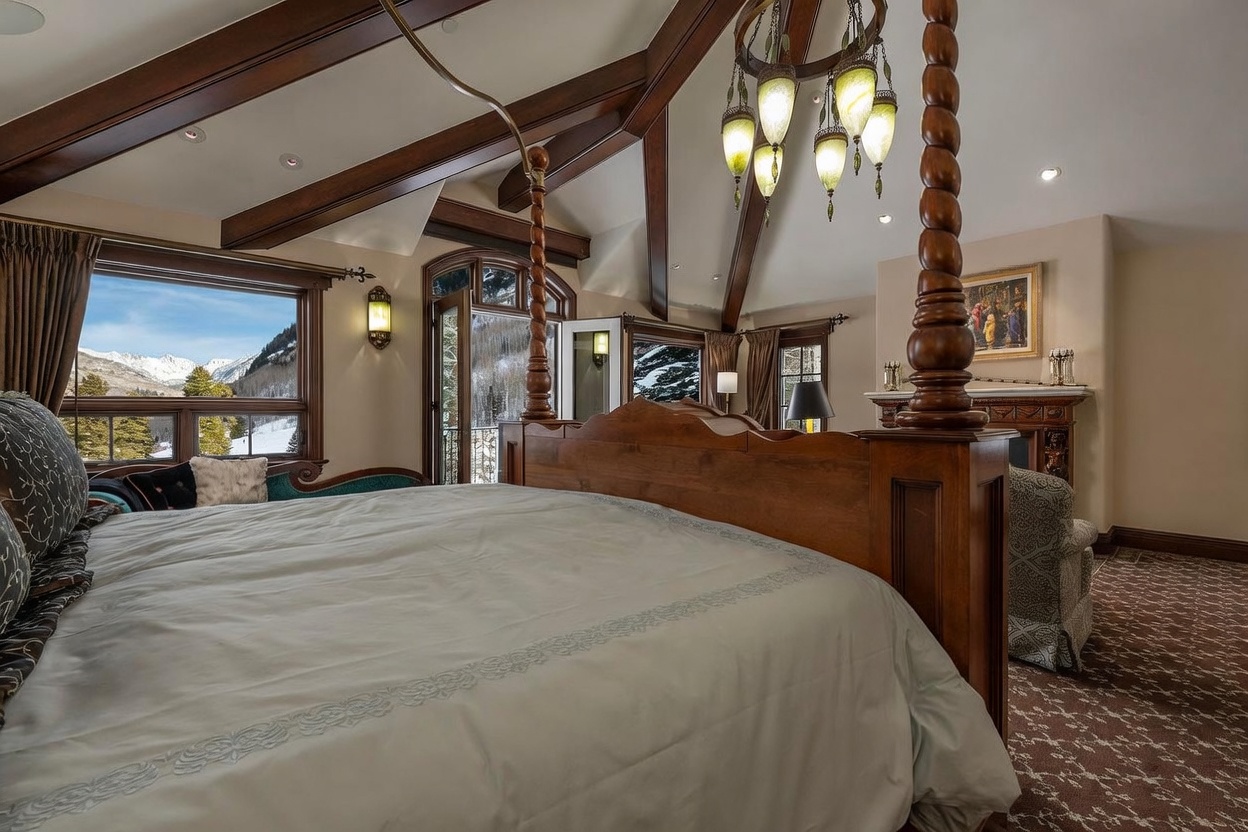 This primary bedroom features a grand four-poster wooden bed as the central focal point, set beneath a vaulted ceiling with exposed dark wood beams. A unique multi-pendant chandelier hangs above the bed, while large windows provide a stunning, unobstructed view of snow-capped mountains. The room includes a cozy seating area near a fireplace and a private balcony access, creating a luxurious and serene mountain retreat atmosphere.
