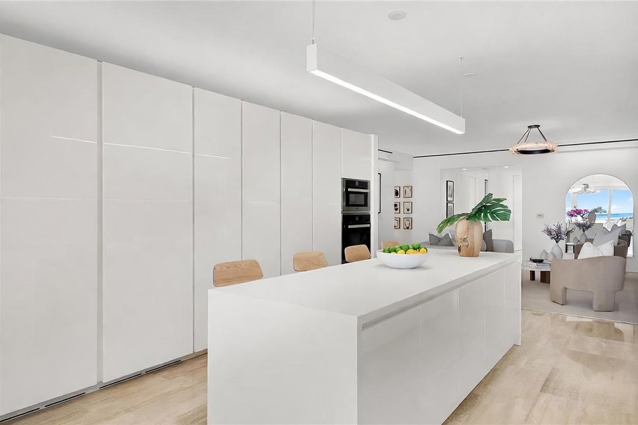 This bright, modern kitchen features sleek white cabinetry and a large central island with light wood stools, creating a minimalist and airy aesthetic. The space is illuminated by a long, linear pendant light, and the open-concept design flows seamlessly into a living area with a view of the ocean through a large arched opening. The light-toned flooring and clean lines contribute to a sophisticated, coastal-inspired atmosphere.