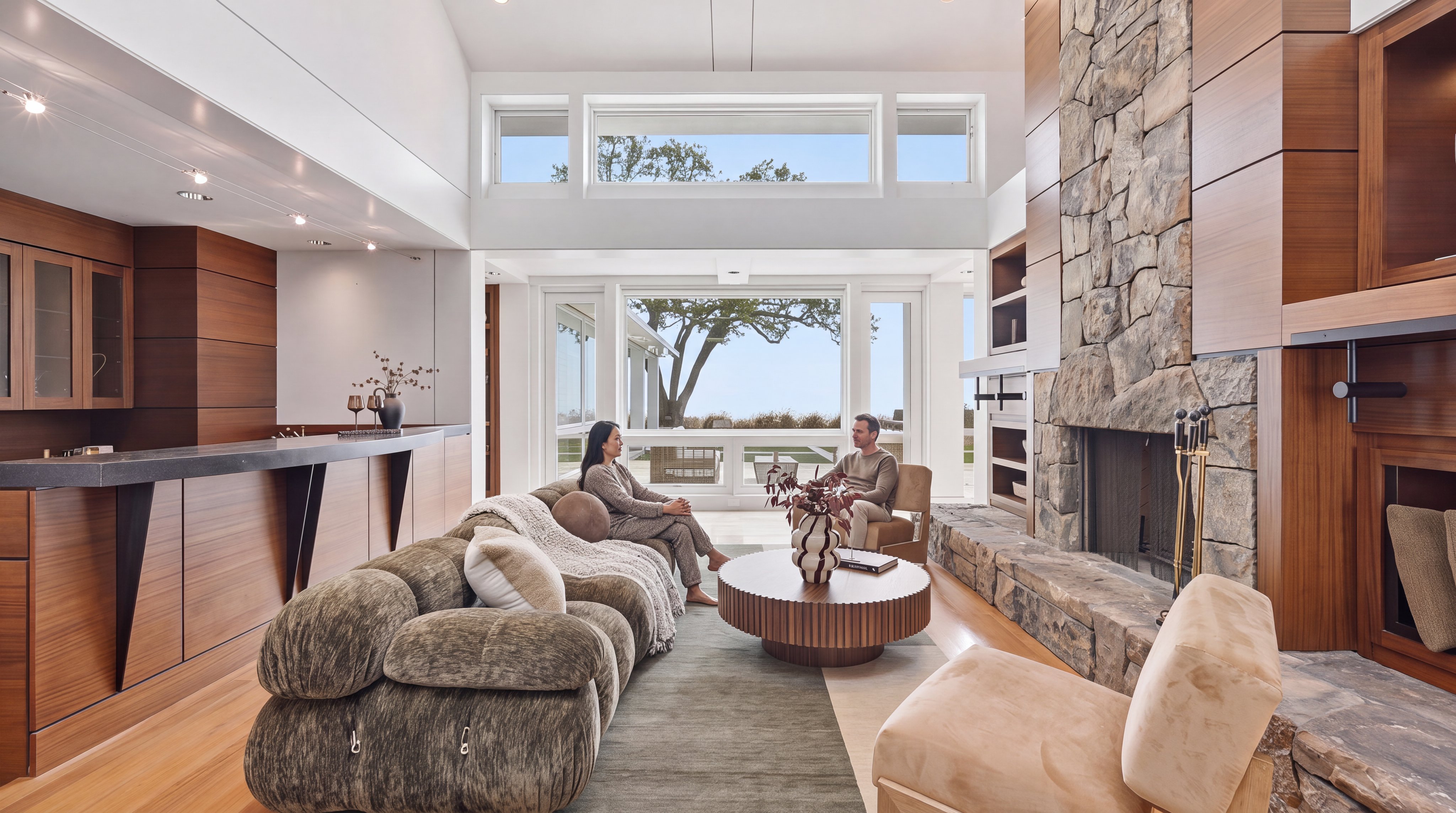 This spacious, modern open-concept living room features high vaulted ceilings, expansive windows that offer panoramic outdoor views, and a prominent floor-to-ceiling stone fireplace. The room is warmly furnished with a plush textured sectional, a circular wooden coffee table, and an intimate seating arrangement, creating an inviting atmosphere that blends contemporary luxury with natural textures.