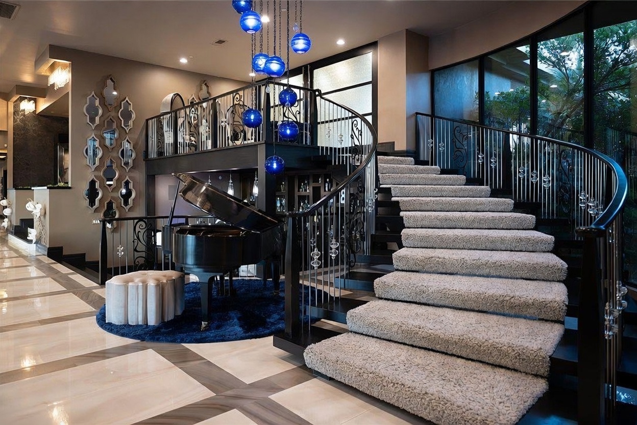 This grand foyer features a sweeping, carpeted staircase with a decorative metal railing that serves as the focal point of the space. A polished black grand piano sits beneath a striking cluster of blue glass pendant lights, resting on a circular blue rug that contrasts with the light-toned, patterned tile flooring. The area is accented by a wall of ornate, decorative mirrors and large windows that provide a view of the exterior greenery, creating an elegant and sophisticated atmosphere.