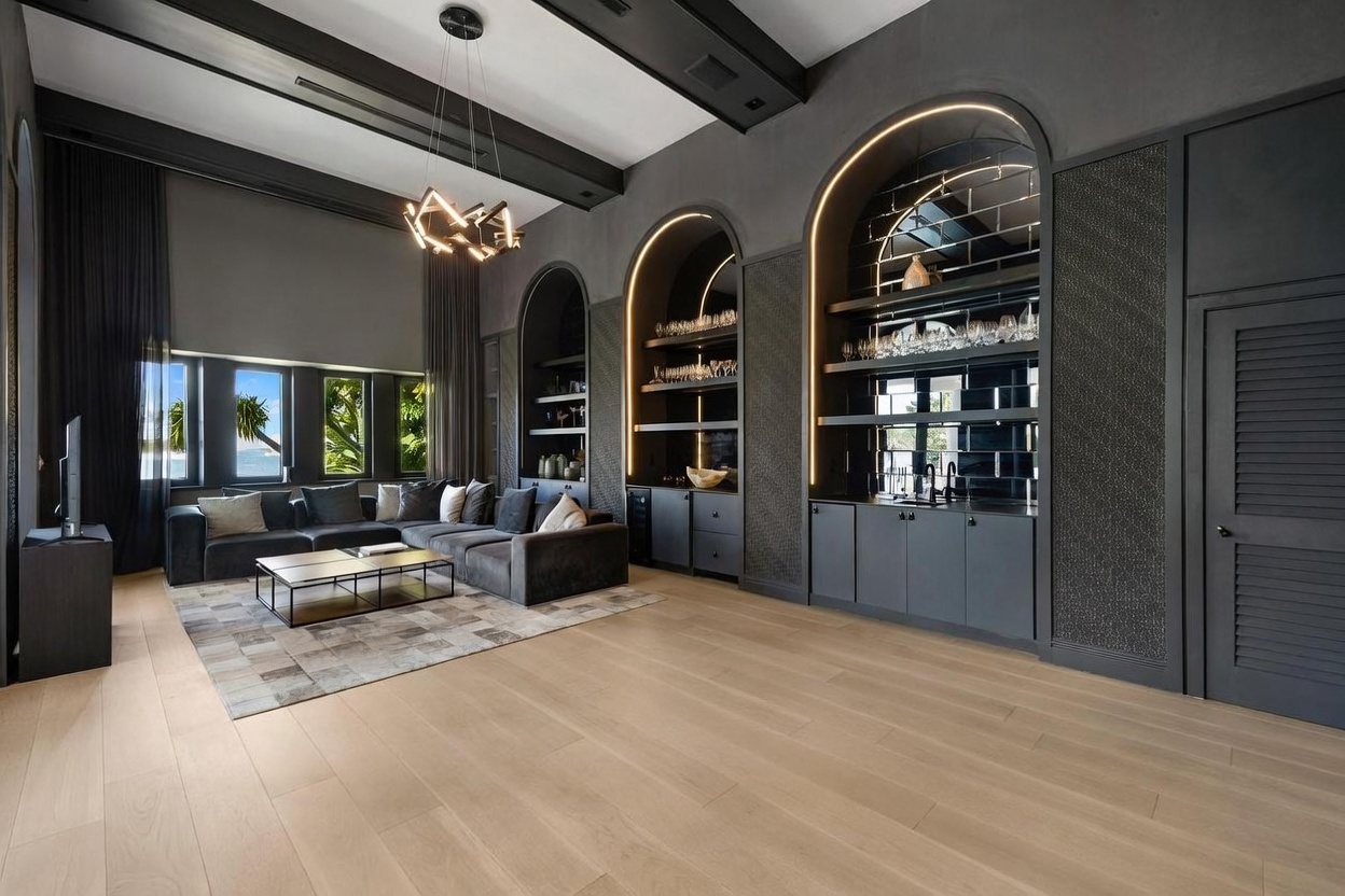 This sophisticated living room features a dark, moody color palette contrasted by light wood flooring and a geometric chandelier. The space includes a large, plush sectional sofa arranged around a low-profile coffee table, set against a backdrop of arched, illuminated built-in shelving and a wet bar. Large windows with dark curtains allow natural light to filter in, creating a dramatic and luxurious atmosphere.