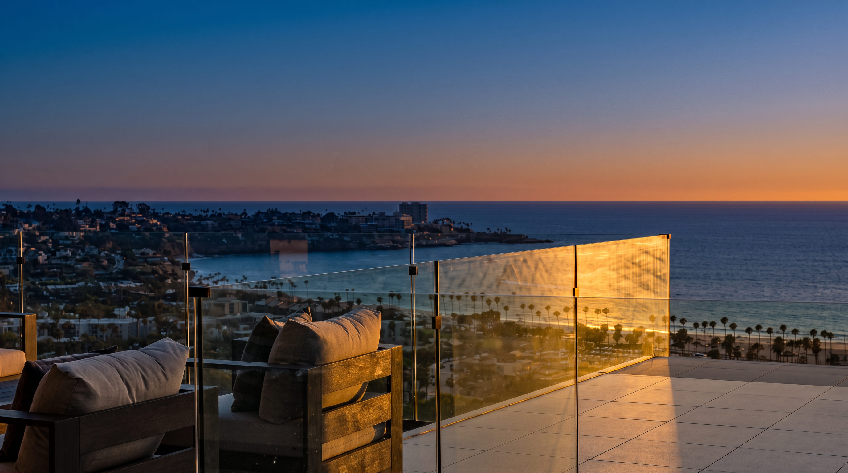 This elevated balcony offers a breathtaking, unobstructed panoramic view of the coastline, ocean, and surrounding residential area. The space is furnished with comfortable outdoor seating featuring plush cushions, set against a modern glass railing that maximizes the scenic vista. The perspective is from a high vantage point, emphasizing the luxurious and serene atmosphere of this outdoor living space.