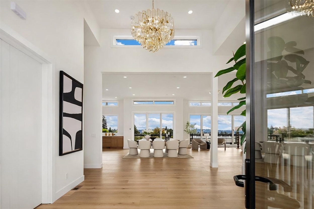 This perspective captures a grand entryway looking through to a bright, open-concept dining area. The space features light wood flooring, a dramatic tiered crystal chandelier, and tall windows that frame a scenic outdoor view. The clean, white walls and minimalist decor create an airy, sophisticated atmosphere that emphasizes the home's spacious layout.