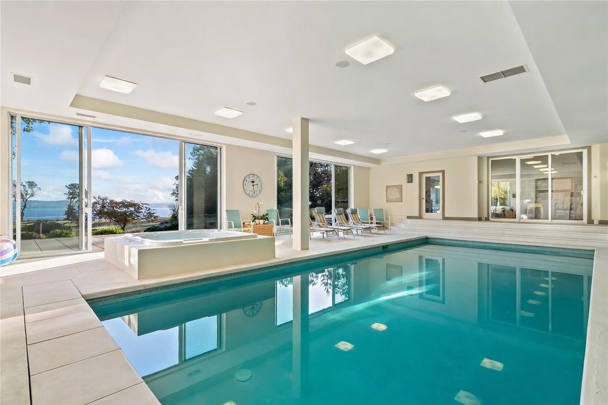 This indoor pool room features a large, rectangular swimming pool adjacent to a raised, built-in spa. The space is surrounded by light-colored tile flooring and multiple lounge chairs, with expansive glass sliding doors providing a panoramic view of the outdoor landscape and water. The room is illuminated by numerous recessed ceiling lights, creating a bright and serene atmosphere.