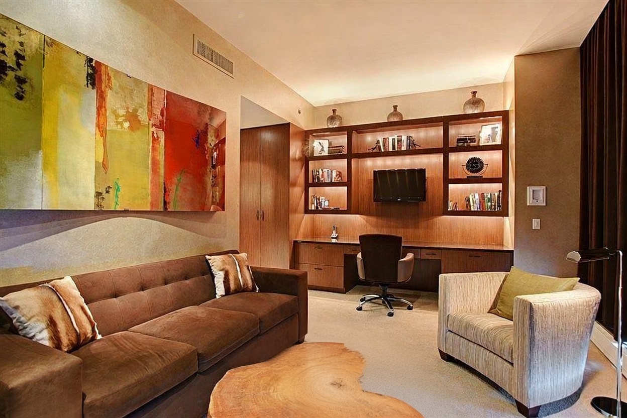 This sophisticated home office and lounge area features a rich, warm color palette with a brown sofa and a matching armchair arranged around a natural wood-slab coffee table. A built-in wood desk unit with integrated shelving and cabinetry provides a dedicated workspace, complete with an ergonomic office chair and a wall-mounted television. The room is accented by a large, vibrant abstract painting on the wall, creating a professional yet comfortable atmosphere suitable for both work and relaxation.