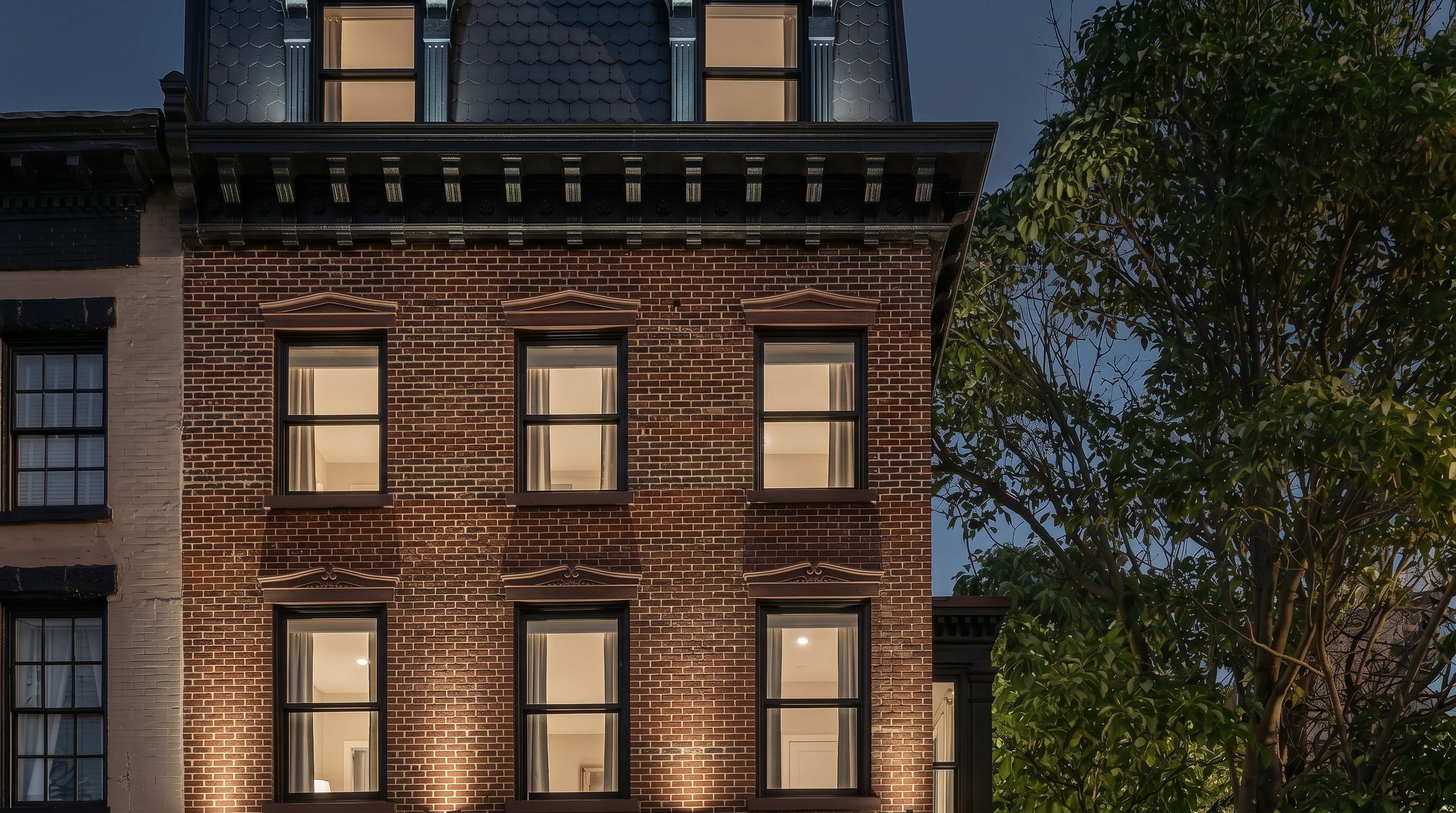 This classic brownstone or brick townhouse facade features traditional architectural details, including a mansard roof with dormer windows and decorative lintels above each sash window. The distinctive red brick work and dark trim accents create an elegant, historic urban aesthetic. A large, mature tree to the right adds a soft element to the structured, formal character of the building.