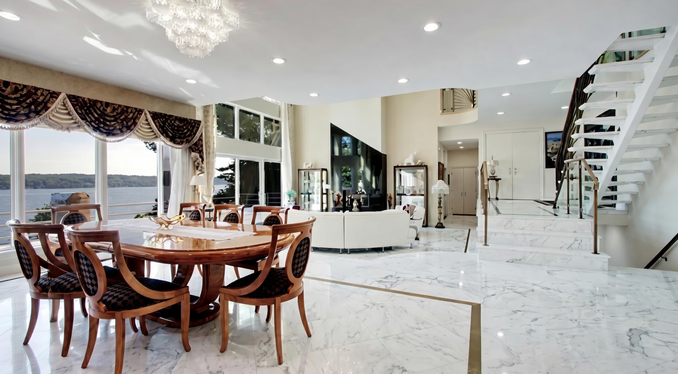 This elegant dining room features a large, dark wood oval table surrounded by matching chairs with patterned upholstery, situated on polished marble floors. A grand crystal chandelier hangs above, and the room connects seamlessly to an open-plan lounge area with a white sectional sofa and sweeping views of the water through floor-to-ceiling windows. The architectural layout includes a striking staircase with a glass railing, contributing to a sophisticated and high-end aesthetic.