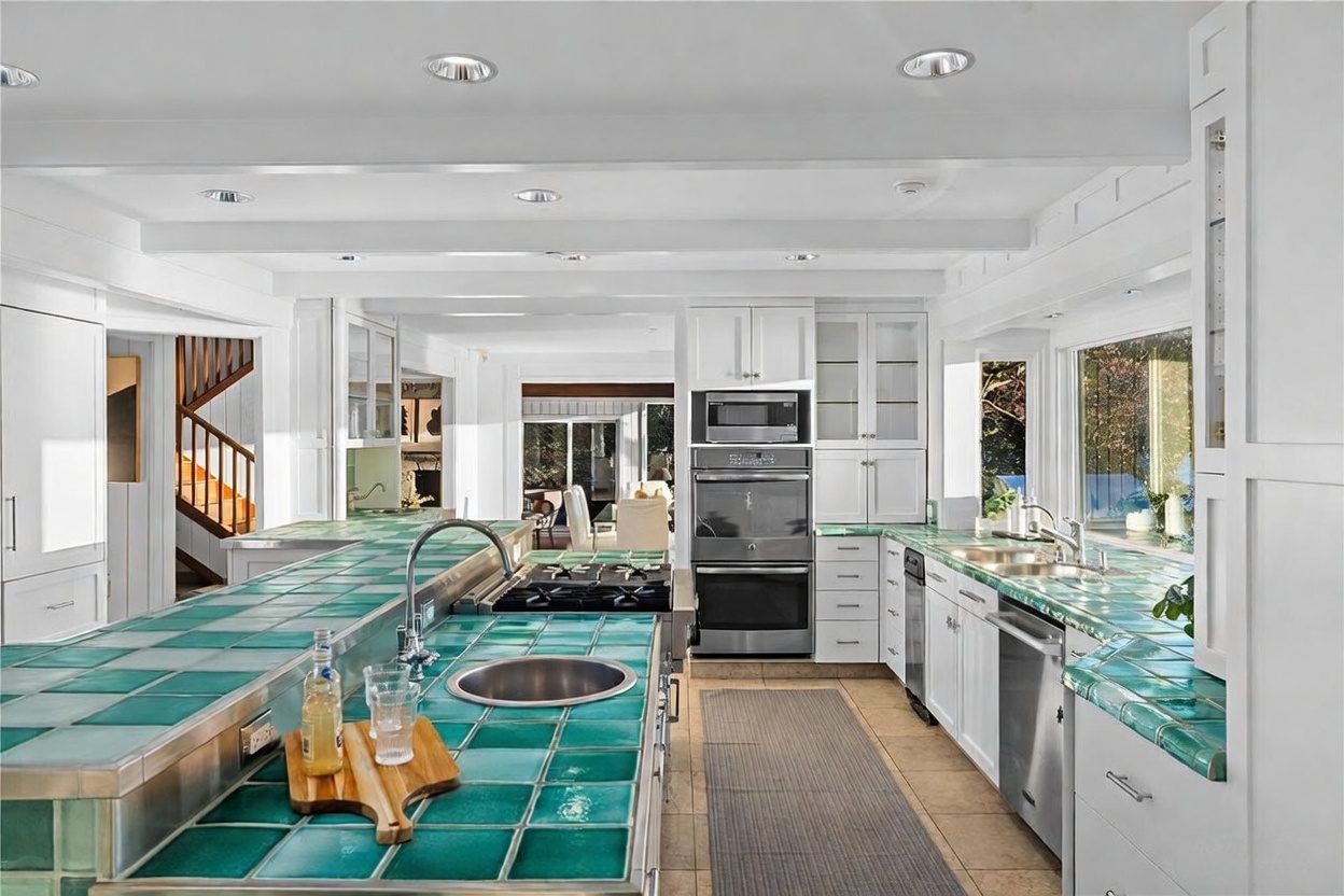 This spacious, bright kitchen features a distinctive large island with a vibrant green tiled countertop and a built-in cooktop. The room is anchored by white cabinetry, stainless steel appliances, and a matching tiled backsplash, creating a unique, retro-inspired aesthetic. The perspective looks directly down the center of the kitchen, highlighting the functional layout and the open transition to the adjacent living area.