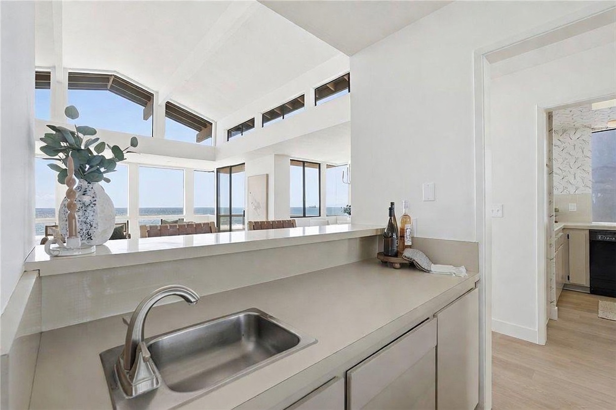 This interior view captures a wet bar or secondary kitchen area featuring a stainless steel sink and light-toned countertops. The space opens up to a bright, airy living area with large windows that provide a clear view of the ocean beyond. The perspective is from behind the counter, looking out toward the expansive, light-filled living space with its vaulted ceiling.