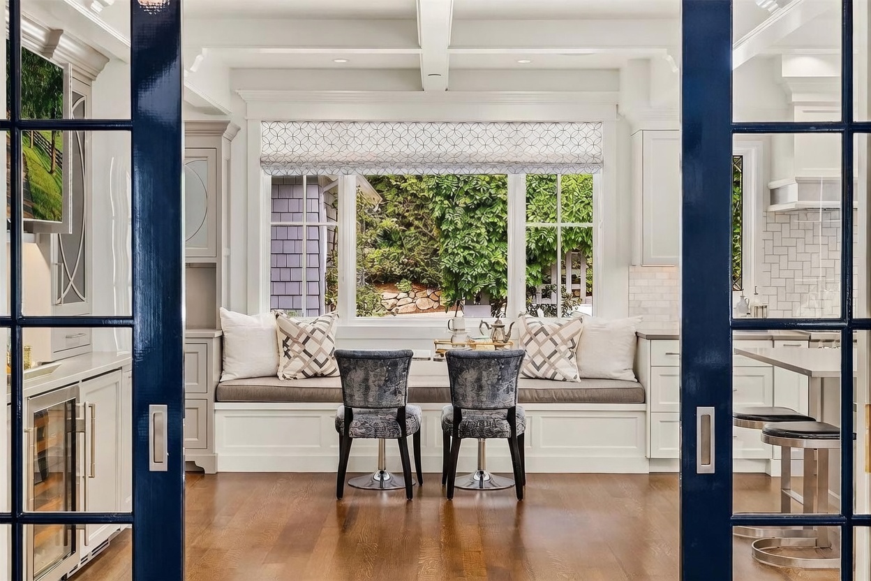 This view captures a bright breakfast nook framed by dark blue French doors, creating a sophisticated transition into the space. The area features a built-in window seat with plush cushions and patterned pillows, centered beneath a large window that overlooks lush greenery. Two upholstered chairs are positioned at the nook, and the overall aesthetic is clean, elegant, and inviting.