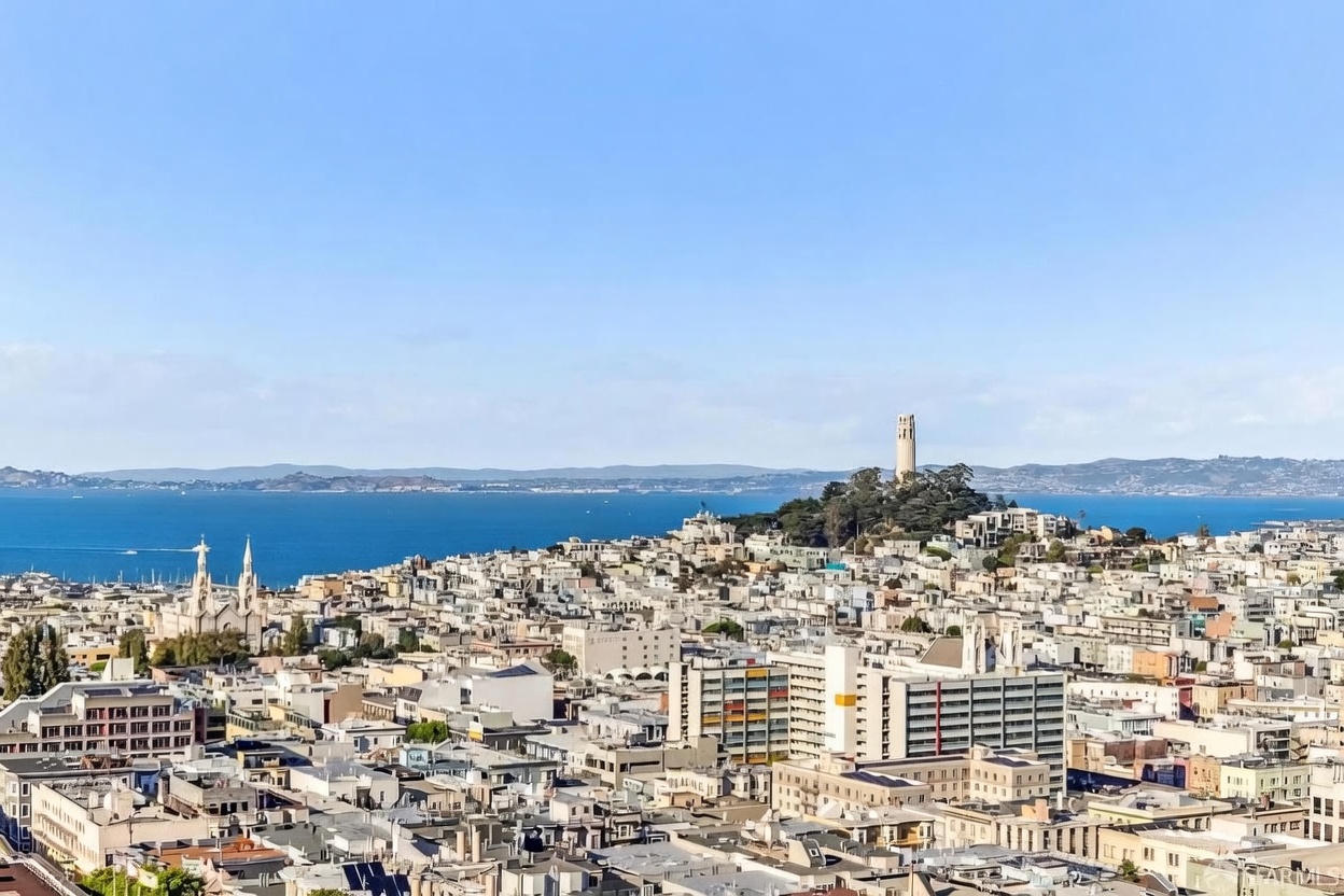 This high-angle aerial view captures a sprawling cityscape of San Francisco, showcasing a dense urban landscape filled with historic architecture and multi-story buildings. In the distance, the iconic Coit Tower stands prominently atop Telegraph Hill, framed by the deep blue waters of the bay and distant hills. The perspective provides a cinematic sense of scale, highlighting the city's unique topography and its proximity to the waterfront.