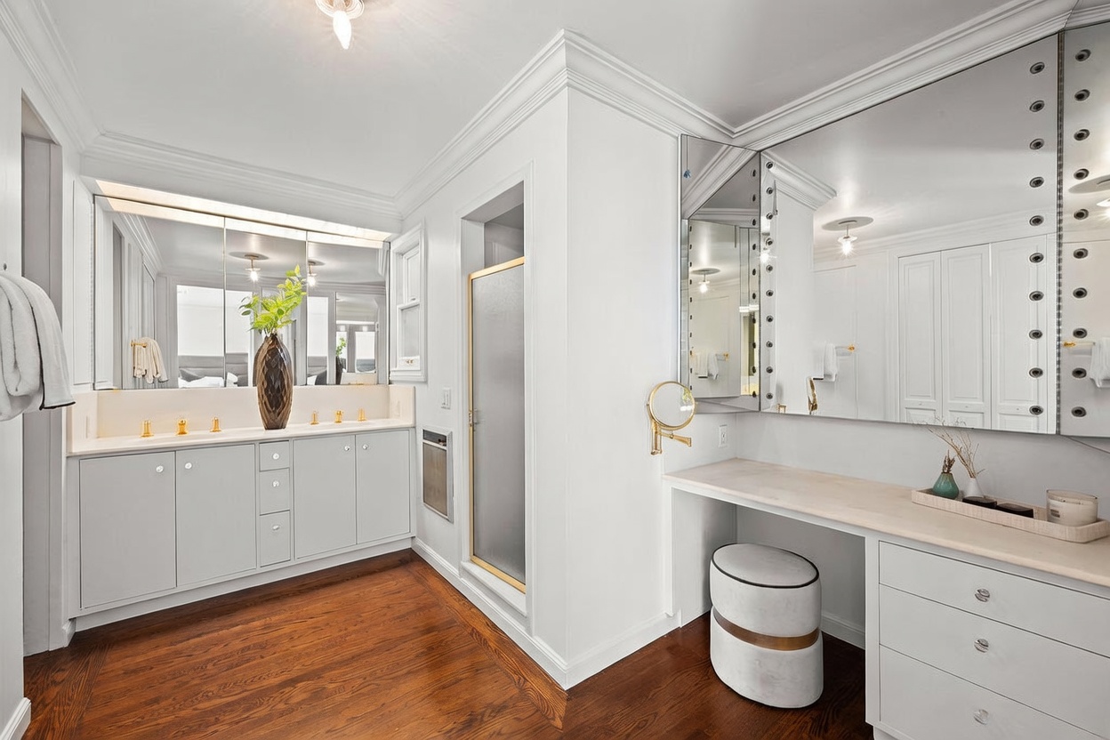 This bright and elegant primary bathroom features a dual-sink vanity with white cabinetry and a large mirror, complemented by a separate vanity area with a stool. The space includes a glass-enclosed shower and rich wood flooring, creating a clean and sophisticated aesthetic. The perspective captures the full layout of the room, highlighting the ample storage and vanity space.