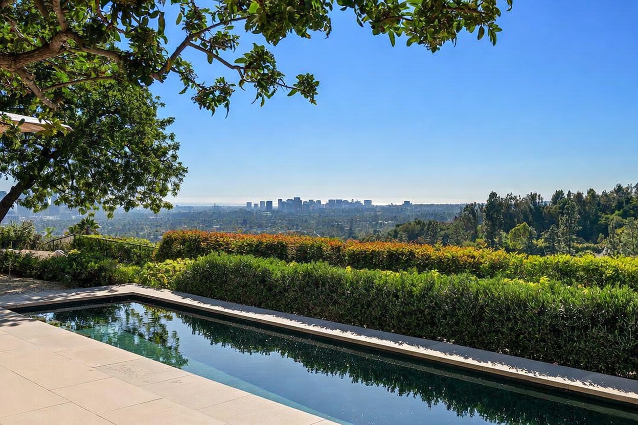 This image captures a serene, rectangular swimming pool set against a breathtaking panoramic view of a distant city skyline. The foreground features a clean, light-colored stone pool deck bordered by a lush, manicured hedge, while a large tree canopy frames the top of the scene. The perspective is at eye level, emphasizing the seamless transition between the private luxury of the pool area and the expansive urban landscape beyond.