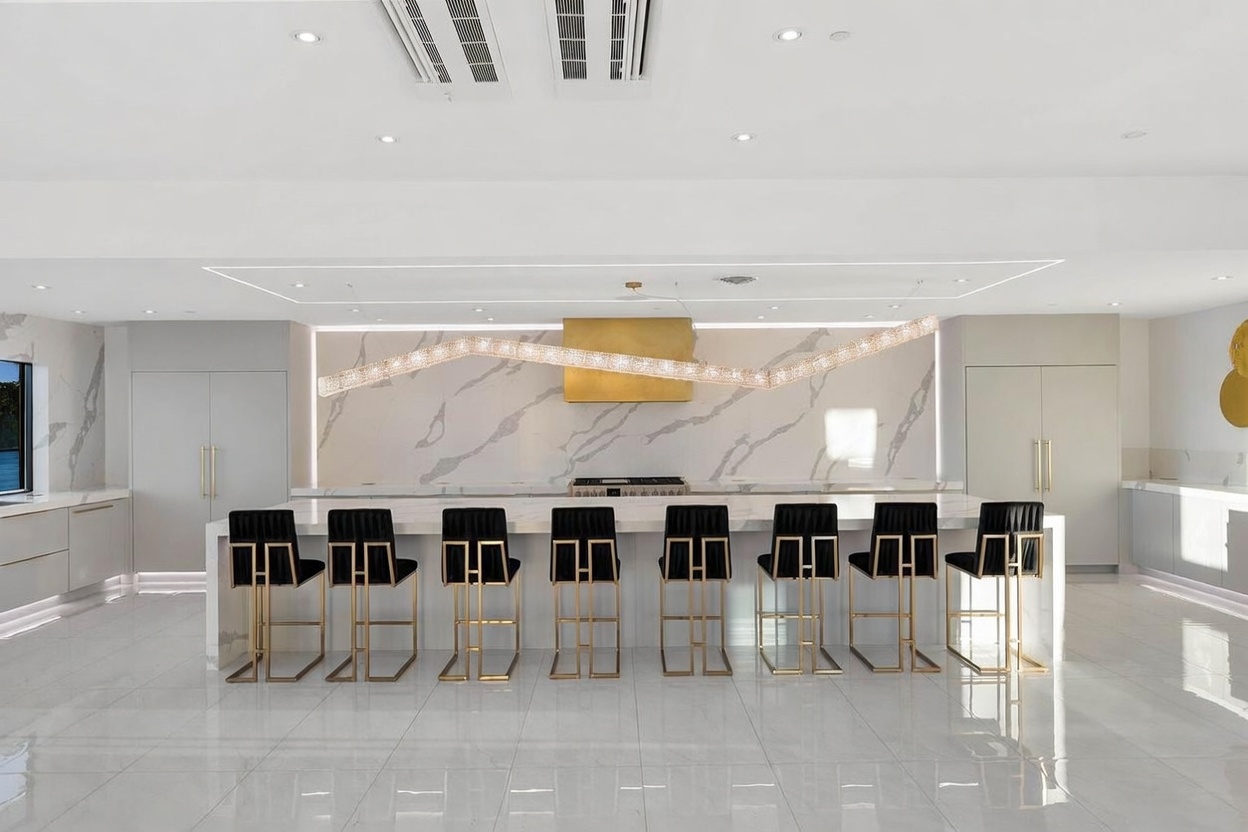 This ultra-modern kitchen features a massive central island with seating for eight, highlighted by a striking, undulating linear chandelier. The space is defined by a sleek, minimalist aesthetic with a large stone-clad backsplash, a gold-toned range hood, and high-gloss white cabinetry. The perspective is a wide, symmetrical shot that emphasizes the expansive, polished flooring and the clean, contemporary lines of the room.