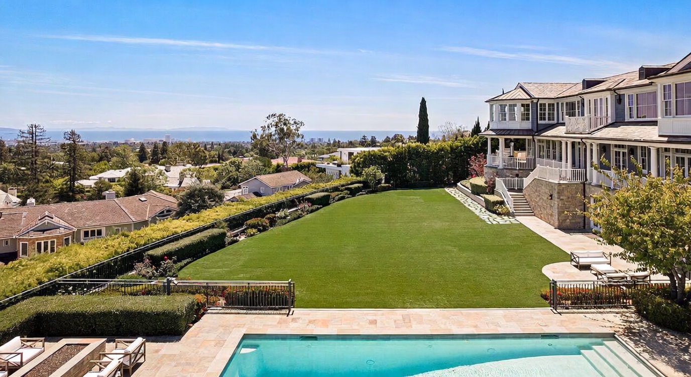 This elevated rear view captures a sprawling estate featuring a large, manicured lawn bordered by lush hedges and mature trees. A stone-paved patio surrounds a rectangular swimming pool in the foreground, while the multi-story residence showcases traditional architectural details and a spacious balcony. The expansive horizon offers a scenic view of the distant coastline and city landscape, creating a serene and luxurious atmosphere.