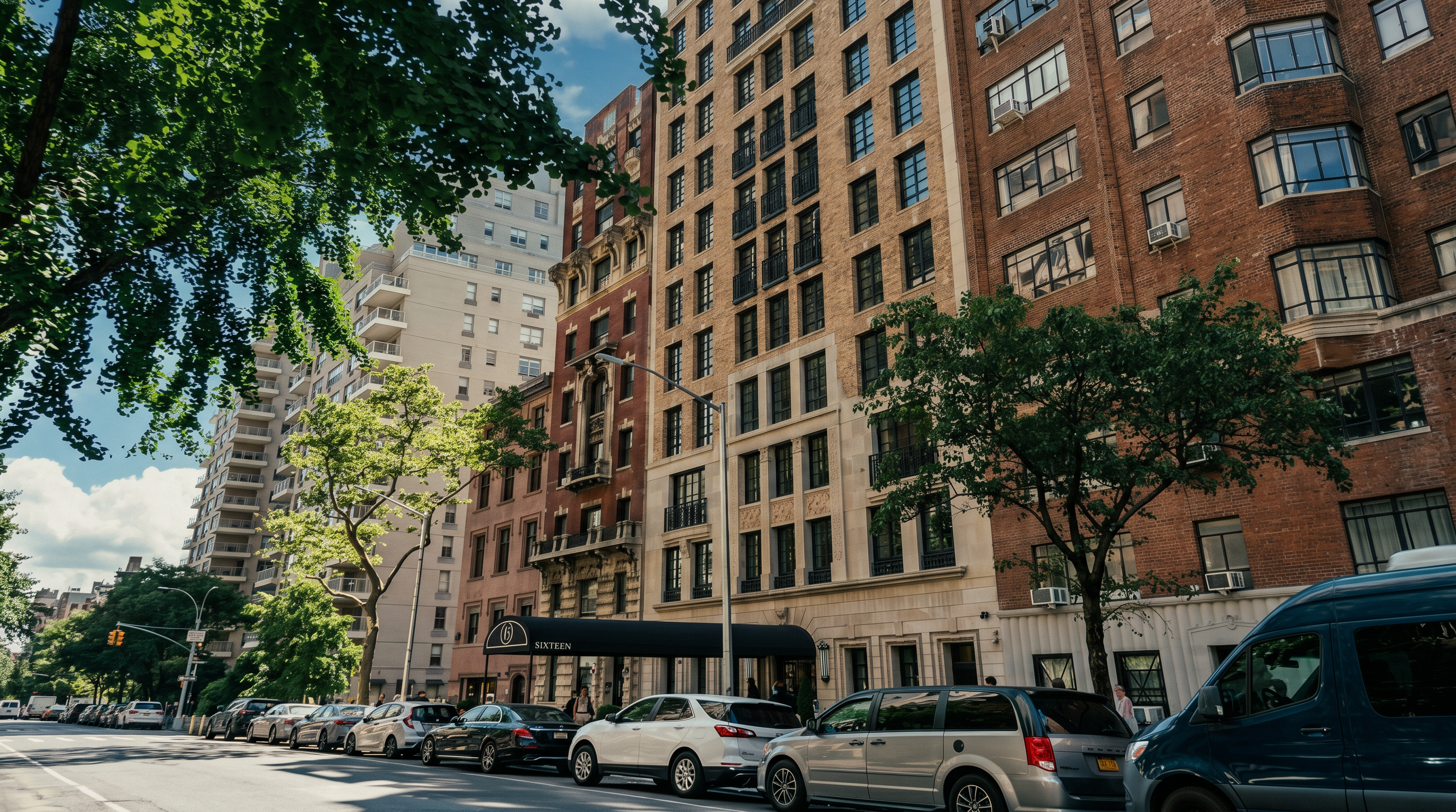 This low-angle, street-level perspective captures the grand facade of a classic, multi-story brick apartment building in a dense urban setting. The architecture features a mix of traditional masonry, symmetrical window arrangements, and a prominent black awning marking the building's entrance. Lush green trees frame the foreground, contrasting with the structured, vertical lines of the building and the parked cars along the street.