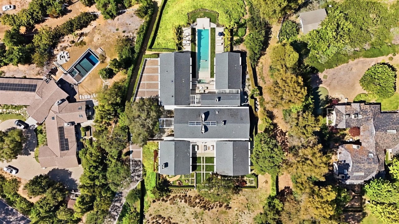 This high-altitude aerial view captures a sprawling, multi-winged estate surrounded by lush, mature landscaping and dense trees. The property features a prominent rectangular swimming pool centered in the rear courtyard, flanked by symmetrical building wings and manicured green spaces. A long, winding driveway leads to the front of the residence, showcasing the expansive scale and private, secluded nature of the grounds.