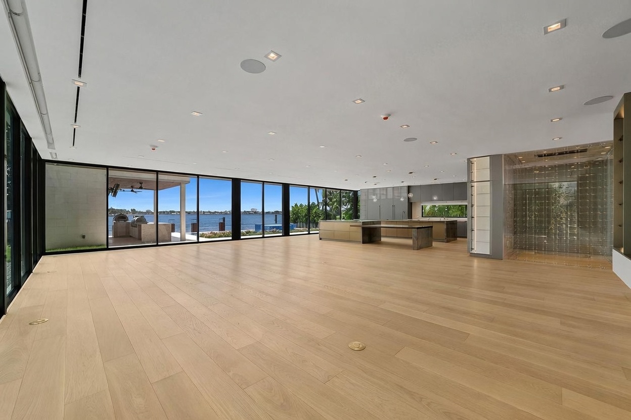 This expansive, open-concept living area features light wood flooring and a wall of floor-to-ceiling glass panels that provide an unobstructed view of the waterfront. To the right, a modern kitchen with a large island and a glass-enclosed wine display creates a sophisticated, minimalist aesthetic. The room is illuminated by numerous recessed ceiling lights, emphasizing the vast, airy feel of the space.