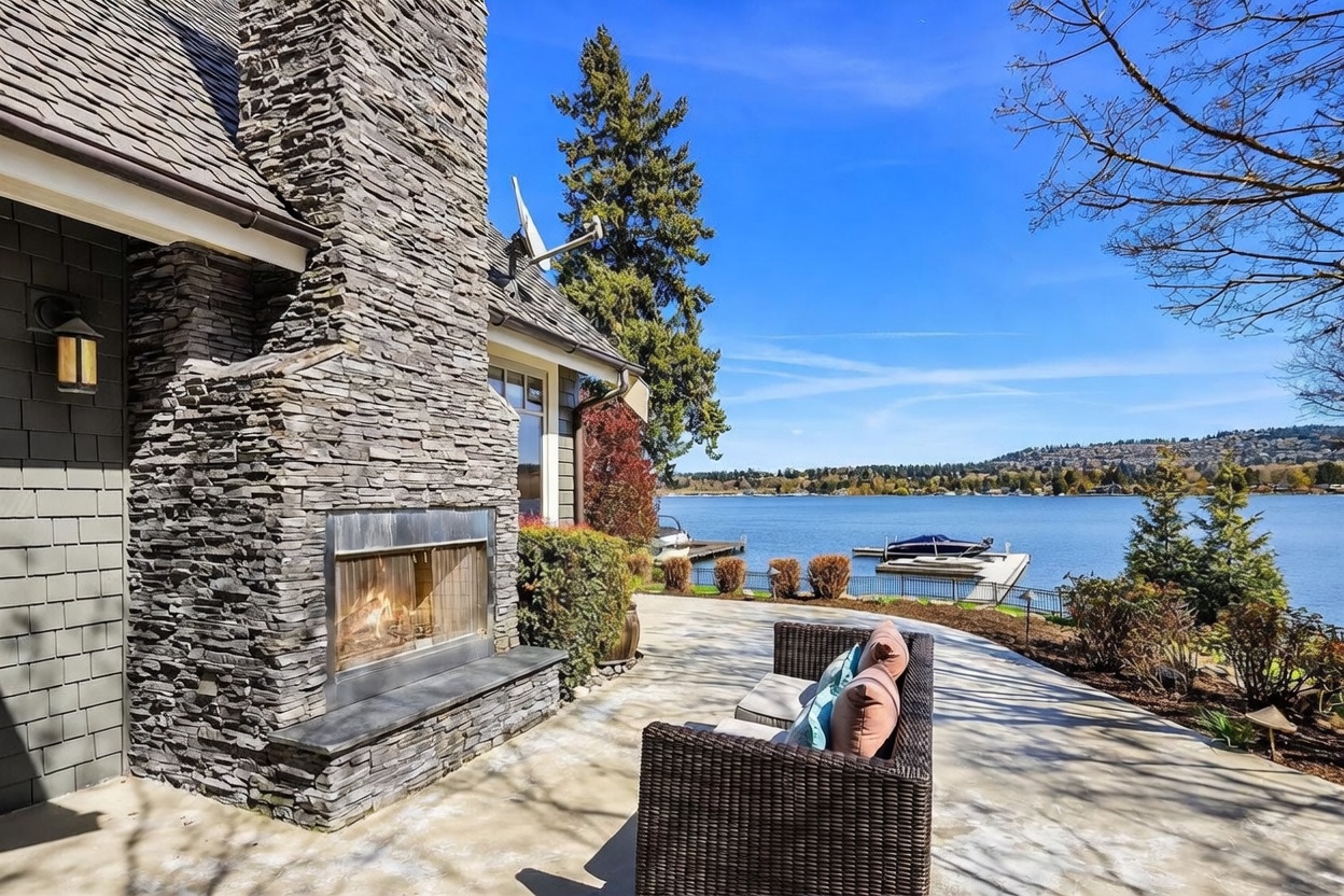 This serene waterfront patio features a prominent stone-clad outdoor fireplace, creating a cozy focal point against the expansive lake backdrop. The space is furnished with comfortable wicker seating, perfect for enjoying the panoramic views of the water and distant shoreline. The scene captures a peaceful, high-end outdoor living experience with a clear blue sky overhead.