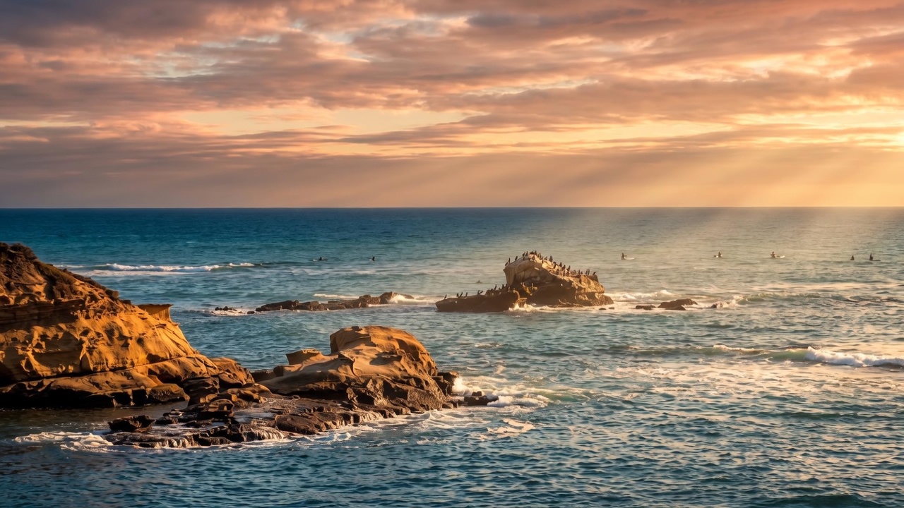 This scenic coastal view captures a rugged shoreline with weathered rock formations jutting out into the deep blue ocean. The perspective is elevated, looking down over the water where small waves break against the cliffs and a distant rock island populated by birds. The scene conveys a serene, natural atmosphere typical of a prime coastal property location.