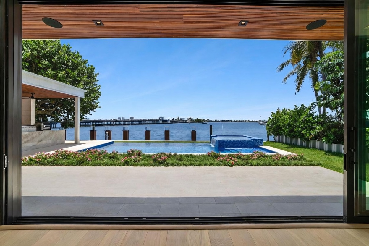 This view is captured from inside a home looking out through large, open sliding glass doors toward a waterfront backyard. The scene features a rectangular swimming pool with an integrated spa, set against a backdrop of a calm bay and a distant bridge. A manicured lawn, vibrant pink flowers, and a covered patio area with a wood-paneled ceiling complete this luxurious outdoor living space.