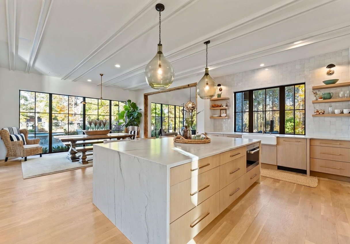 This bright, open-concept kitchen features a large central island with a stone waterfall countertop and light wood cabinetry. The space is illuminated by two prominent glass pendant lights and large windows that provide a view of the adjacent dining area and outdoor scenery. The design combines modern elements with warm, natural tones, creating an inviting and airy atmosphere.