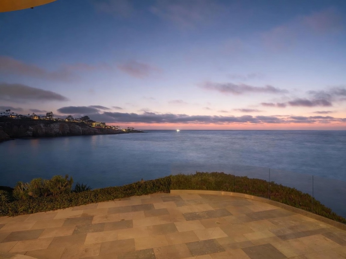 This image captures a spacious stone-paved patio overlooking a serene coastal scene at twilight. A sleek glass railing provides an unobstructed view of the ocean, cliffs, and a soft, colorful sunset sky. The perspective is from an elevated vantage point, emphasizing the tranquil and expansive outdoor living space.