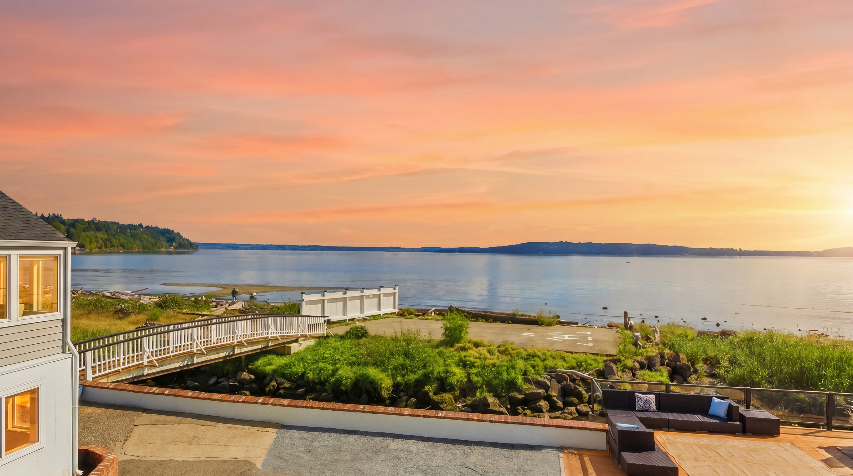 This elevated perspective captures a scenic waterfront property featuring a spacious deck with comfortable outdoor seating. A white pedestrian bridge leads toward a private concrete landing area near the shoreline, which is framed by natural rocky terrain and lush greenery. The expansive view of the calm bay and distant hills creates a serene and inviting atmosphere, perfect for outdoor relaxation.