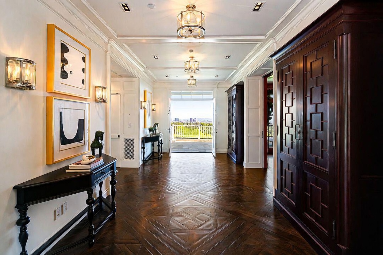 This elegant, wide hallway features rich, dark wood flooring laid in a classic herringbone pattern, leading the eye toward an open doorway that reveals a scenic outdoor view. The space is illuminated by multiple tiered glass chandeliers and wall-mounted sconces, creating a sophisticated atmosphere. A large, dark wood armoire stands to the right, while a sleek console table adorned with art and decor sits to the left, emphasizing a refined and symmetrical design.