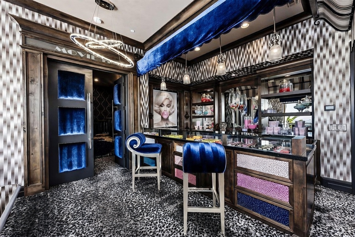 This unique home theater concession area features a vibrant, high-contrast design with a leopard-print carpet and a custom candy bar with glass-fronted display cases. The space is accented by deep blue velvet upholstery on the bar stools and a matching valance, complemented by a portrait of Marilyn Monroe and modern circular pendant lighting. The perspective captures the bar area from a slightly elevated angle, highlighting the transition into the adjacent theater room through a dark, paneled doorway.