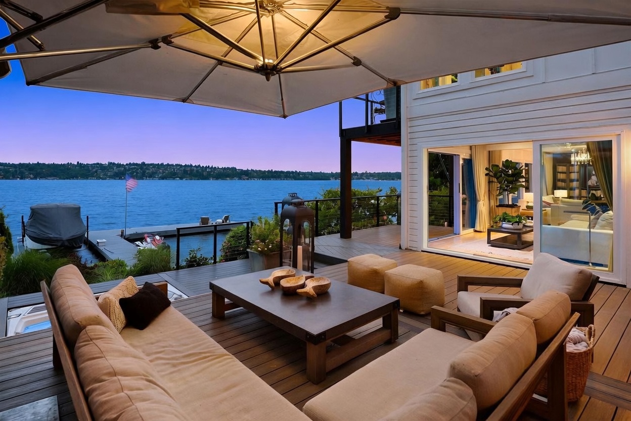 This expansive waterfront deck features a comfortable outdoor lounge area with plush seating, a central coffee table, and two ottomans, all sheltered by a large cantilevered umbrella. The deck overlooks a serene lake with a private dock and boat lift, creating a tranquil and luxurious atmosphere. Large sliding glass doors provide a seamless transition to the interior living space, which is visible in the background.