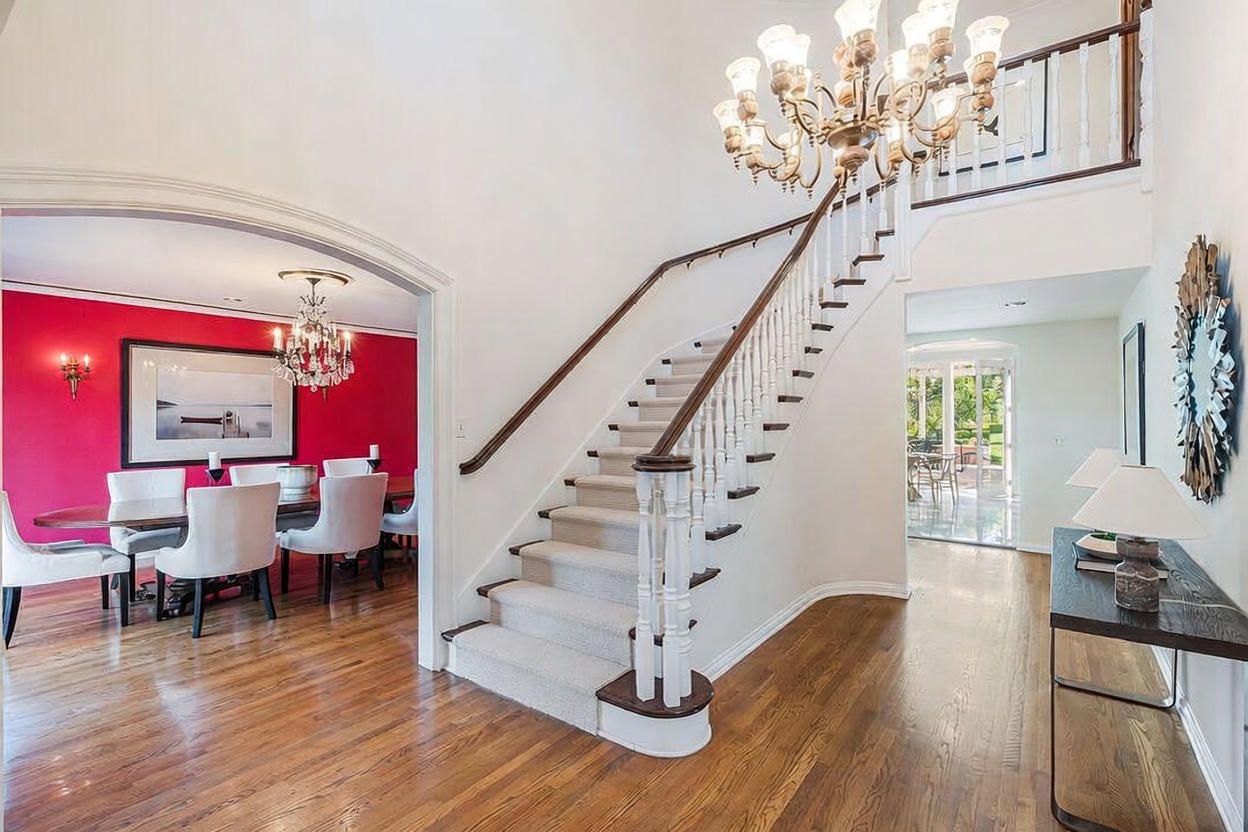 This grand foyer features a prominent central staircase with a light-colored runner and a dark wood handrail, leading up to a landing illuminated by an ornate multi-tiered chandelier. To the left, an arched opening reveals a formal dining room with vibrant red walls and a matching crystal chandelier, while the right side leads toward a bright, open hallway with a console table. The space is characterized by polished wood flooring and a bright, airy atmosphere that creates a welcoming and sophisticated entrance.