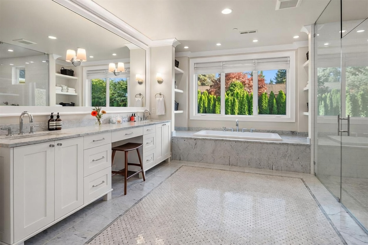 This spacious primary bathroom features a double vanity with stone countertops, a built-in makeup desk, and a large soaking tub positioned beneath a wide window. The room is finished with light-toned cabinetry, a large wall-to-wall mirror, and intricate patterned tile flooring that creates a luxurious, spa-like atmosphere. The perspective is wide-angle, capturing the entire layout from the vanity area across to the glass-enclosed shower on the right.