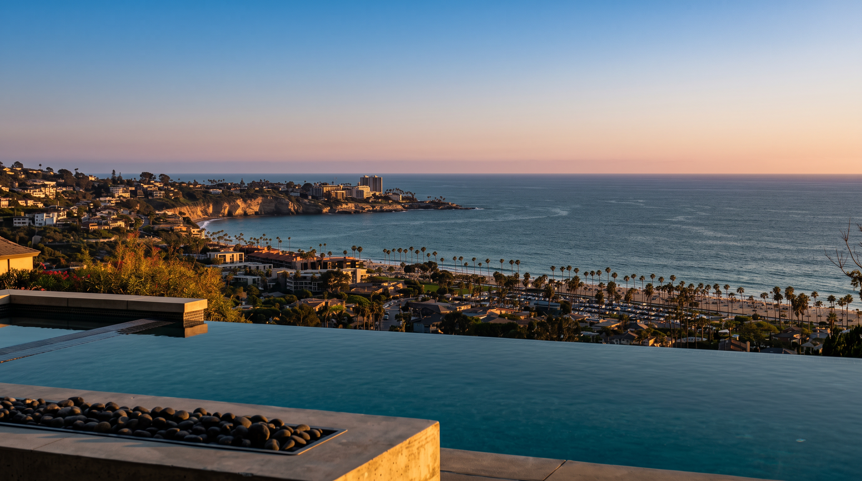 This image captures a stunning infinity-edge pool overlooking a vast coastal landscape and the Pacific Ocean. In the foreground, a stone-lined fire feature sits adjacent to the pool, creating a sophisticated outdoor living space. The perspective is elevated, showcasing a panoramic view of the beach, palm trees, and residential coastline under a clear blue sky.