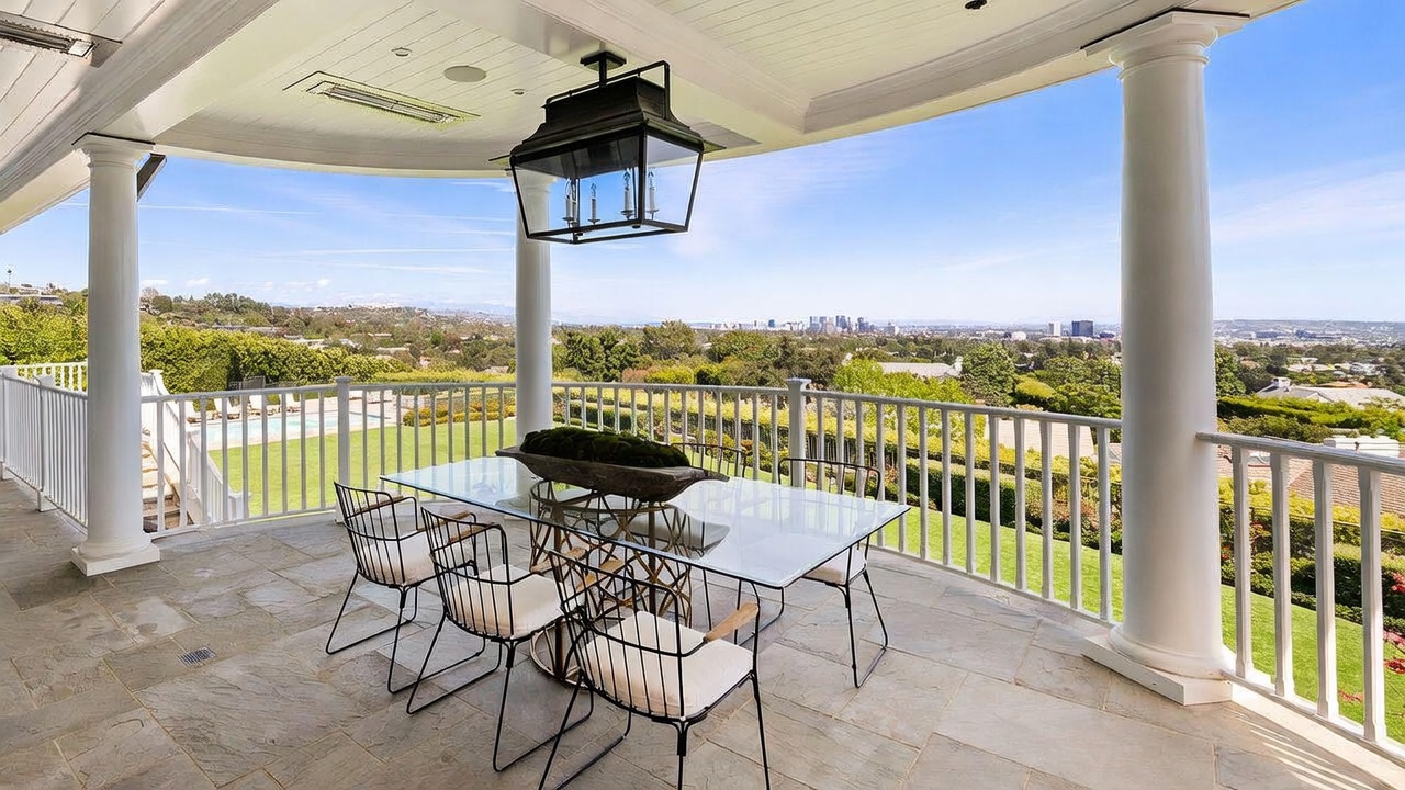 This expansive covered balcony features stone flooring and a white railing, providing a sophisticated outdoor dining area with a glass-topped table and modern wire-frame chairs. A large, dark-framed lantern hangs from the ceiling, adding a touch of elegance to the space. The balcony offers a panoramic, elevated view of the surrounding landscape and distant city skyline, creating an ideal setting for al fresco entertaining.