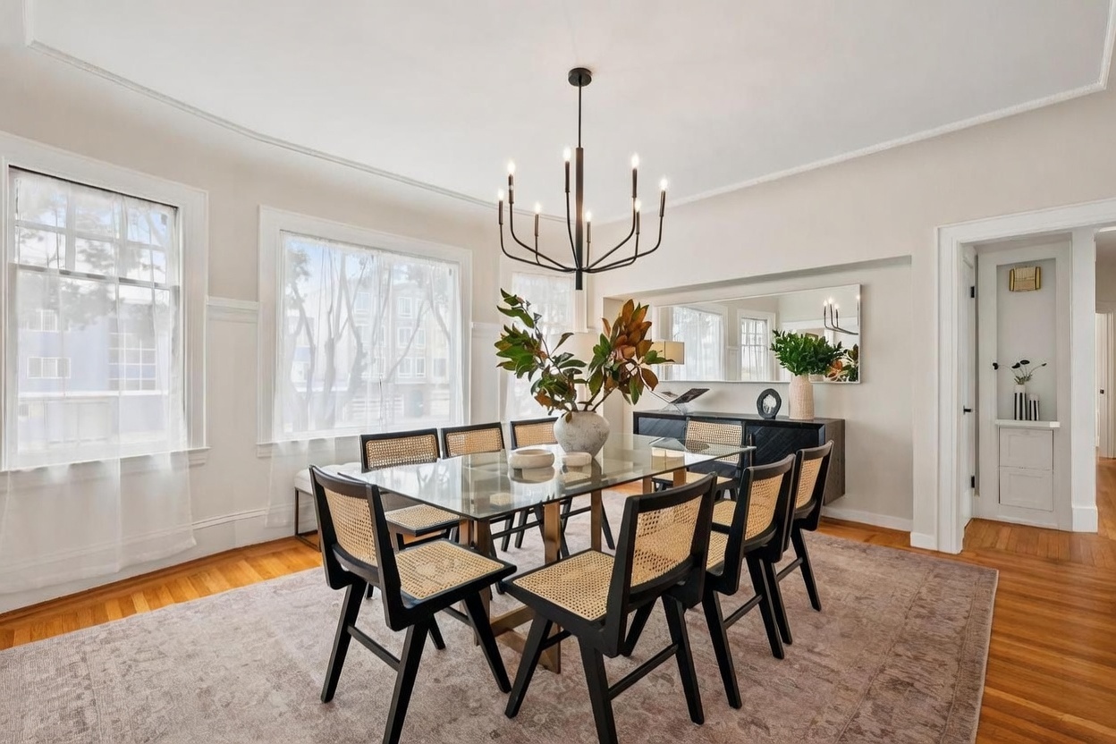 This elegant dining room features a glass-topped table surrounded by black chairs with woven cane detailing, set atop a large, neutral-toned area rug. A modern, multi-armed chandelier serves as a focal point above the table, while a dark sideboard sits against the wall beneath a wide mirror. The space is brightened by large windows with sheer curtains, creating an airy and sophisticated atmosphere.
