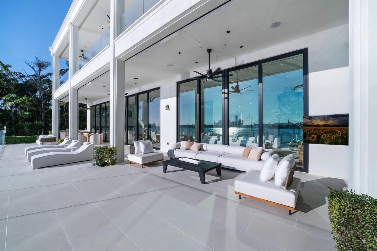 This expansive, modern outdoor patio features sleek white tile flooring and a large, comfortable seating area with a low-profile sofa and a dark coffee table. The space is defined by floor-to-ceiling glass walls that provide a seamless transition to the interior and offer panoramic views of the waterfront and city skyline. A ceiling fan and recessed lighting are integrated into the covered overhang, creating a sophisticated and airy atmosphere perfect for luxury outdoor living.
