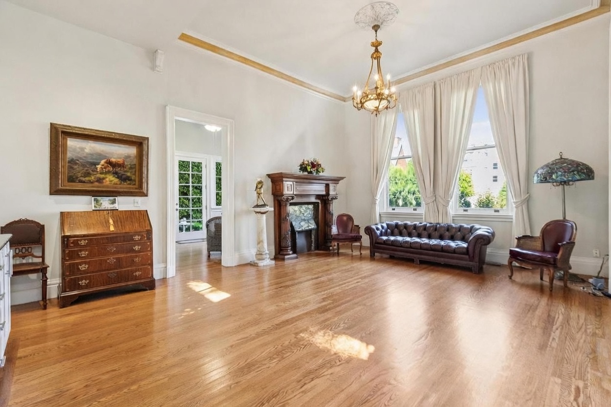 This elegant living room features light-toned walls, polished wood flooring, and a prominent fireplace with a dark wood mantel. A tufted leather sofa and matching armchairs are arranged around the room, complemented by a classic chandelier and tall windows draped in floor-length curtains. The space exudes a traditional, sophisticated atmosphere with a desk and artwork adding to the refined aesthetic.