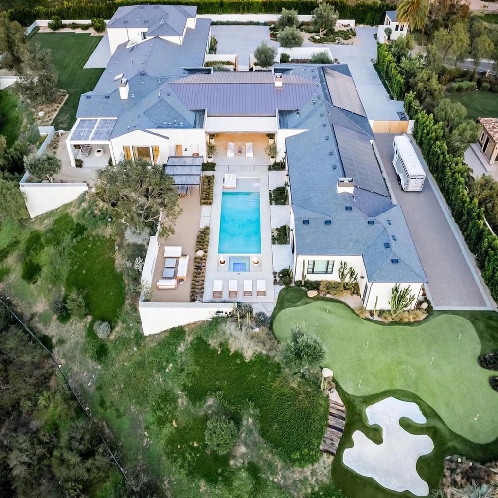 This high-angle aerial view captures a sprawling, modern luxury estate nestled on a hillside. The property features a central swimming pool and spa, a large outdoor patio area with lounge seating, and a private putting green with a sand bunker. The home is characterized by its expansive roofline, solar panels, and seamless integration with the surrounding landscaped grounds and natural terrain.