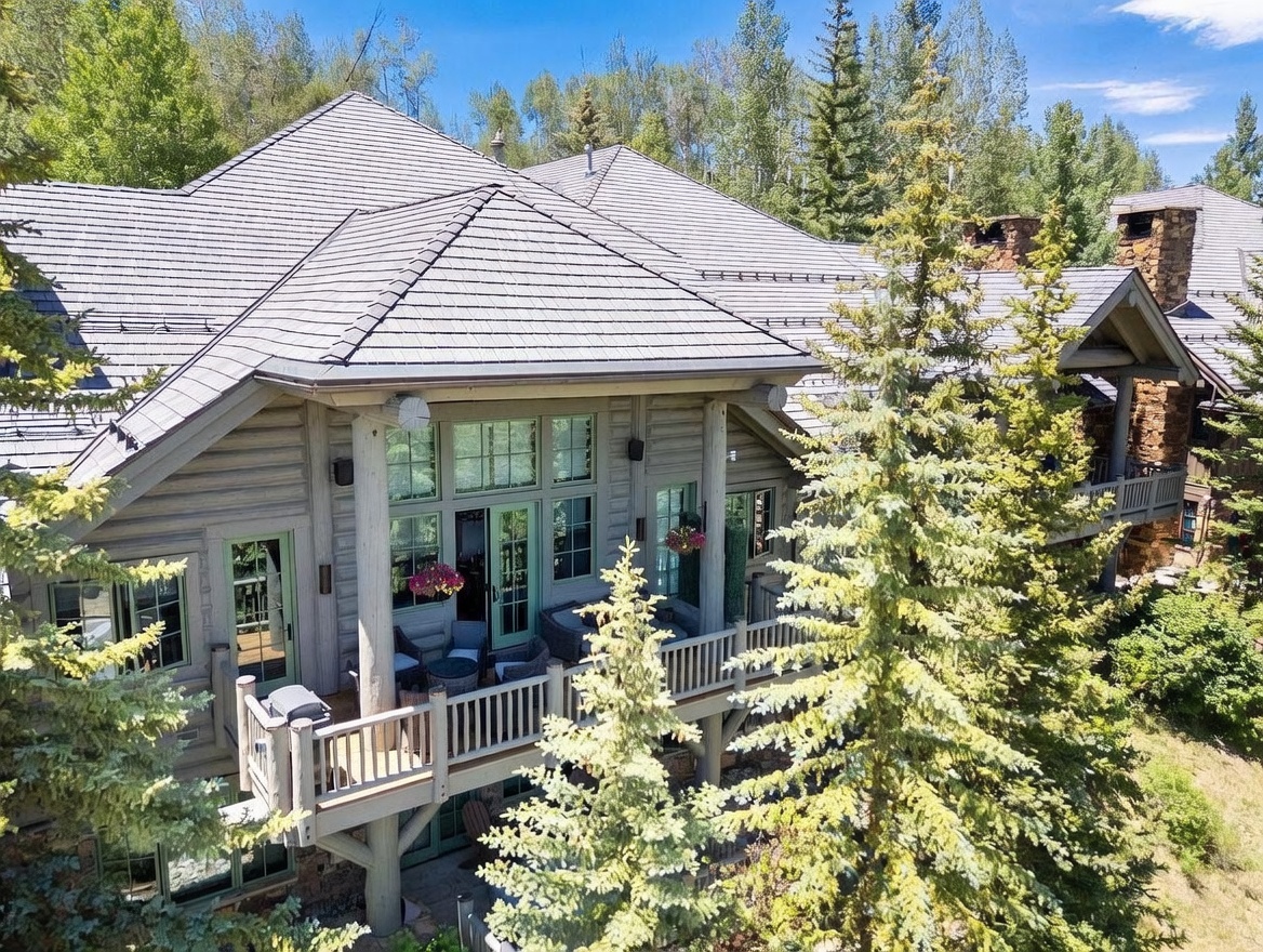 This elevated perspective captures the rear exterior of a rustic log home nestled within a dense, lush forest. The structure features a multi-level wooden deck with outdoor seating, large windows that reflect the surrounding trees, and a complex shingled roofline. The setting is serene and secluded, emphasizing the home's integration into the natural mountain landscape.