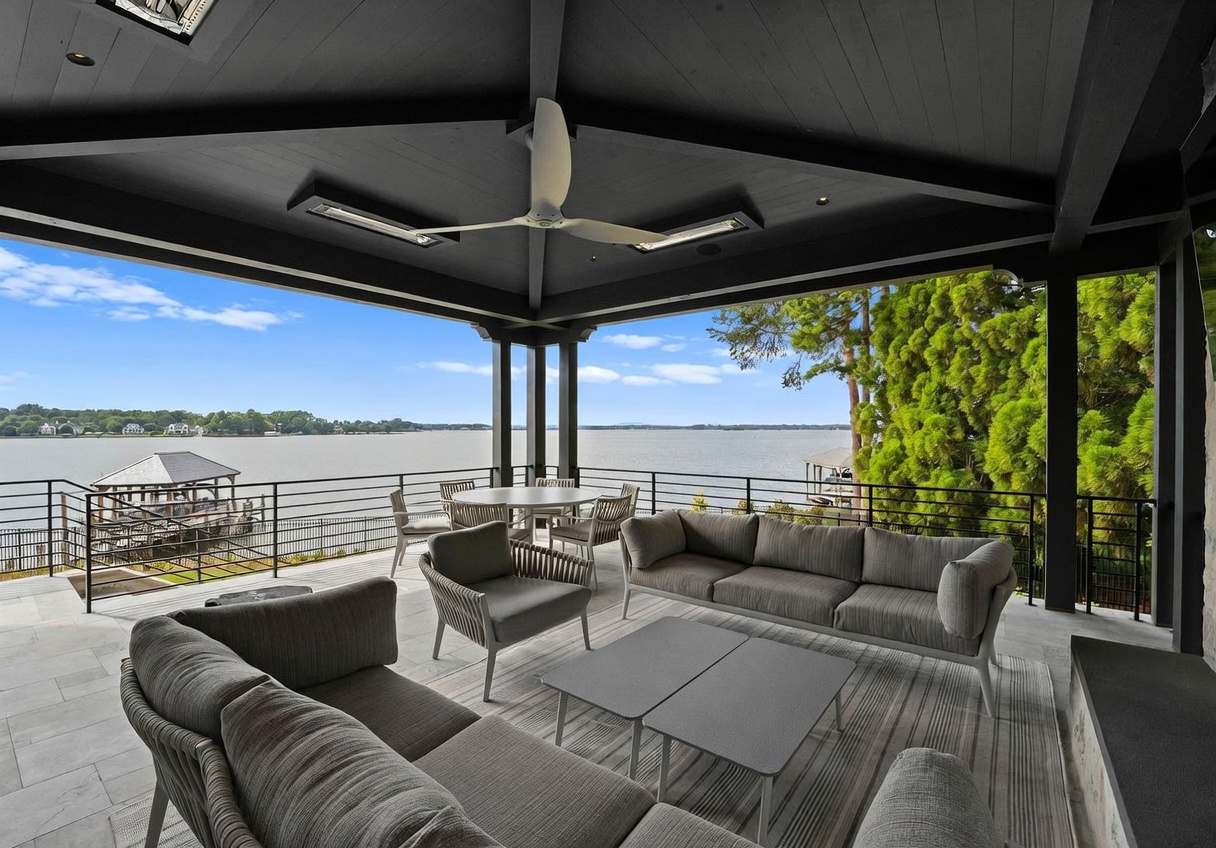 This expansive covered patio offers a sophisticated outdoor living space with panoramic views of the lake. The area is furnished with comfortable grey sectional seating and a central dining set, all sheltered under a dark-toned ceiling equipped with a modern fan and integrated lighting. The sleek metal railing provides safety while maintaining an unobstructed connection to the water and surrounding landscape.
