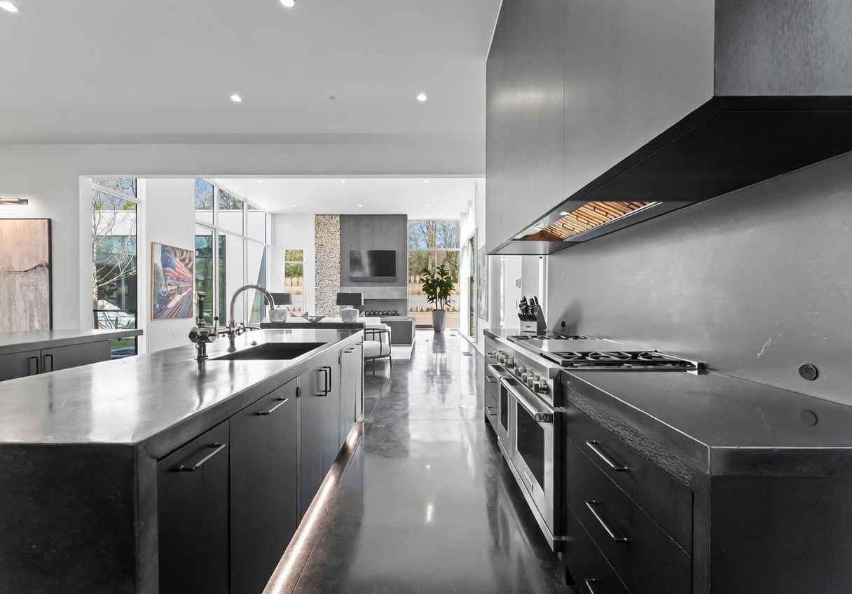 This sleek, modern kitchen features dark cabinetry and a large central island with a dark stone countertop and integrated sink. The space flows seamlessly into an open-concept living area, where large windows provide a view of the outdoor landscape and a tall stone-clad chimney breast. The polished concrete flooring reflects the recessed lighting, creating a sophisticated and cohesive aesthetic throughout the home.
